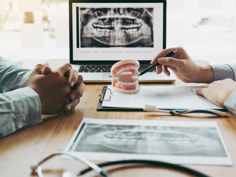 A dental consultation setting with a dentist and a patient discussing a treatment plan. The dentist is holding a pen and pointing at a dental model, while the patient has their hands clasped together. On the table, there is a clipboard with papers, dental X-ray image , a laptop displaying the same X-ray and a dental model.