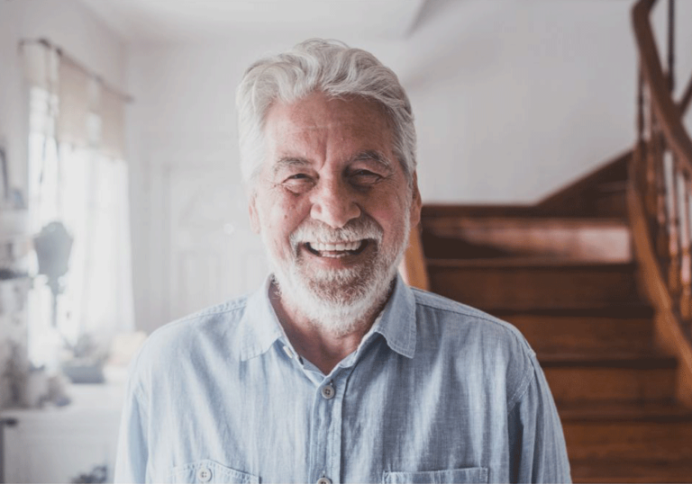 An older individual with short gray hair wears a light blue button-up shirt, standing indoors near a wooden staircase. The background includes sheer curtains and a small decorative plant near a window, creating a cozy, home-like atmosphere.