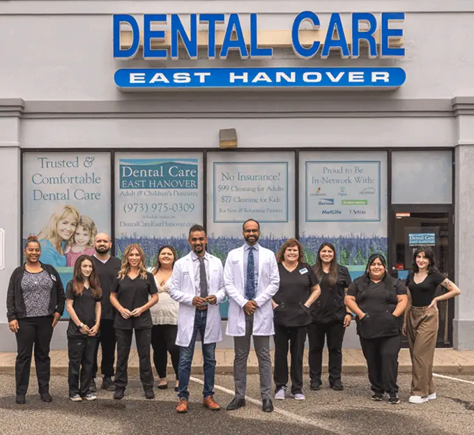 A group of dental care professionals is pictured standing in front of a clinic with signage reading 'Dental Care East Hanover.' The clinic's windows display promotional offers and insurance details, emphasizing accessibility and affordability for patients.